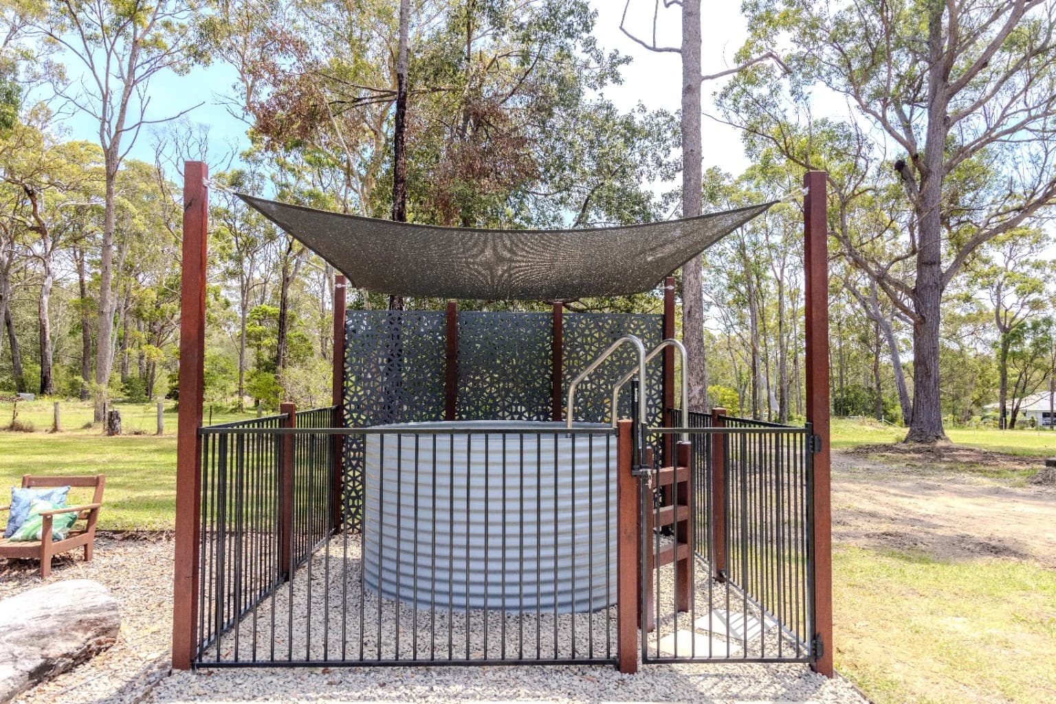 Supplied photograph of a fully above-ground light grey steel pool with shade sail, access ladder and surrounding black fence in a rural garden